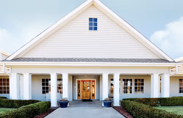 Front exterior view of a single-story building with white siding, a peaked roof, and a covered entrance supported by white columns. There are windows on either side of the wooden double front doors, and neatly trimmed bushes line the walkway leading to the entrance.