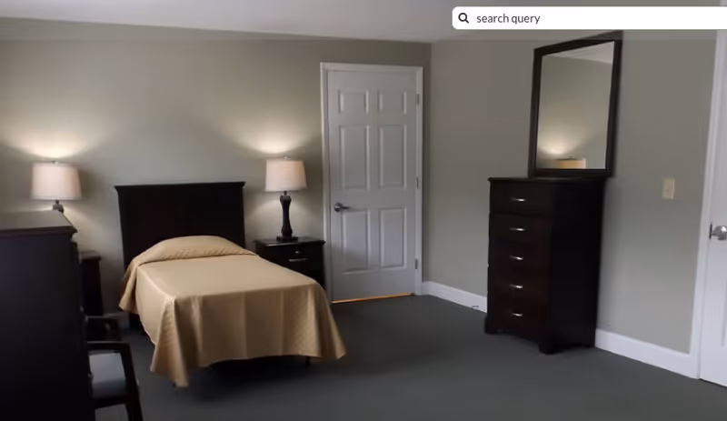 A simple bedroom with a single bed covered with a beige bedspread, two bedside tables each with a lamp, a dark wooden dresser with a mirror, and two closed white doors. The walls are painted light gray and the floor is carpeted in dark green.
