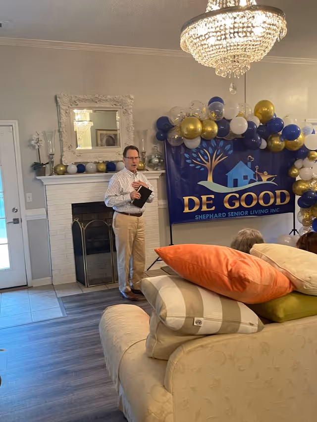 A man in a checkered shirt and beige pants stands near a white brick fireplace with a decorative mirror above it. Behind him is a large banner with balloons that reads 'DE GOOD SHEPHARD SENIOR LIVING INC.' The room has a chandelier, a glass door, and a beige couch with multiple colorful pillows in the foreground.