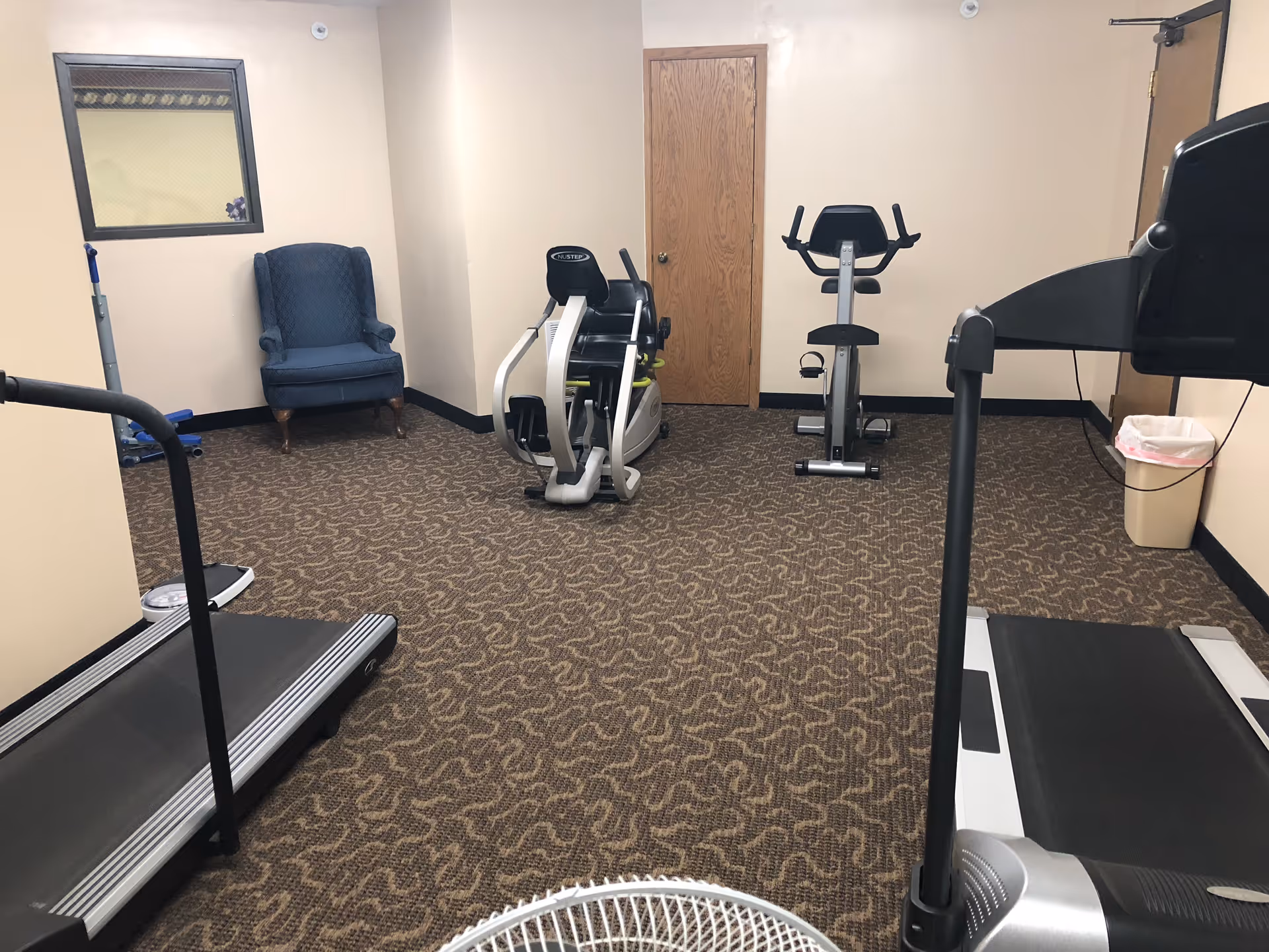 A small exercise room with patterned carpet flooring, beige walls, and a wooden door. The room contains two treadmills, a stationary bike, a recumbent exercise bike, a blue upholstered armchair, a trash can, and a fan in the foreground.