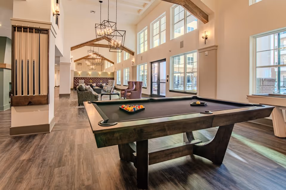A spacious and well-lit recreational room featuring a pool table with balls arranged for a game. The room has large windows allowing natural light to fill the space, wooden beams on the ceiling, modern chandeliers, and comfortable seating areas with sofas and armchairs in the background.