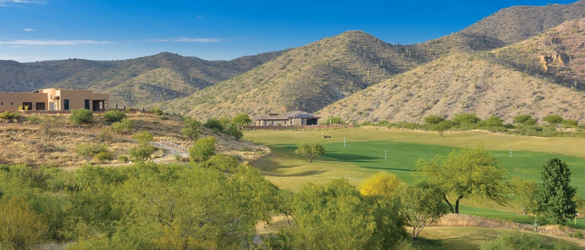 Scenic view of a golf course surrounded by green trees and desert hills with two modern buildings in the background under a clear blue sky.