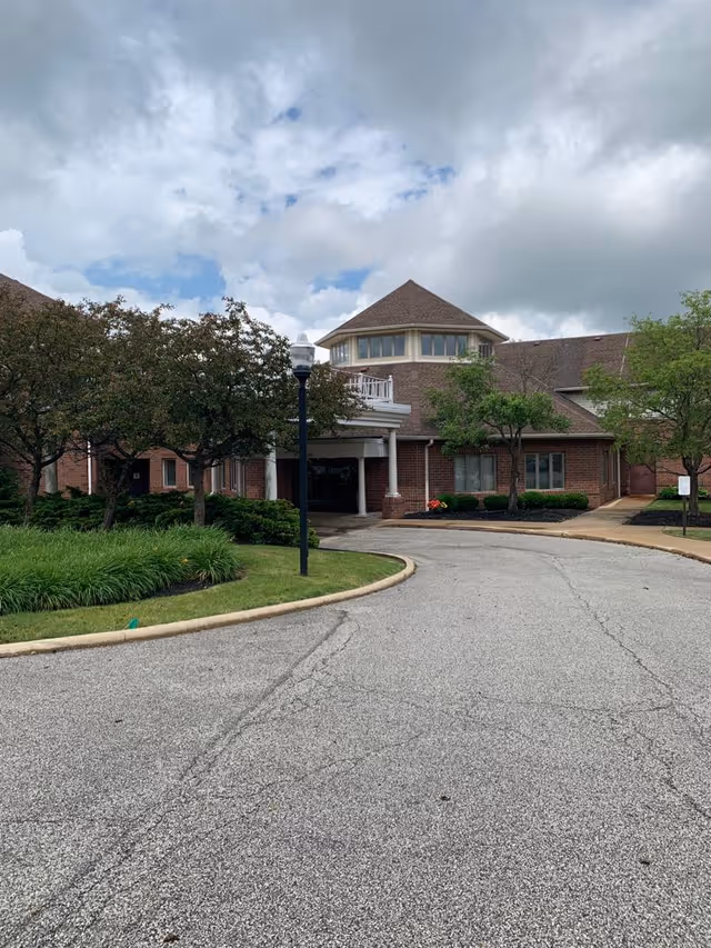Exterior view of a brick building with a peaked roof and multiple windows, surrounded by trees and landscaping. A curved driveway leads to the entrance, which is covered by a white portico supported by columns. The sky is partly cloudy.