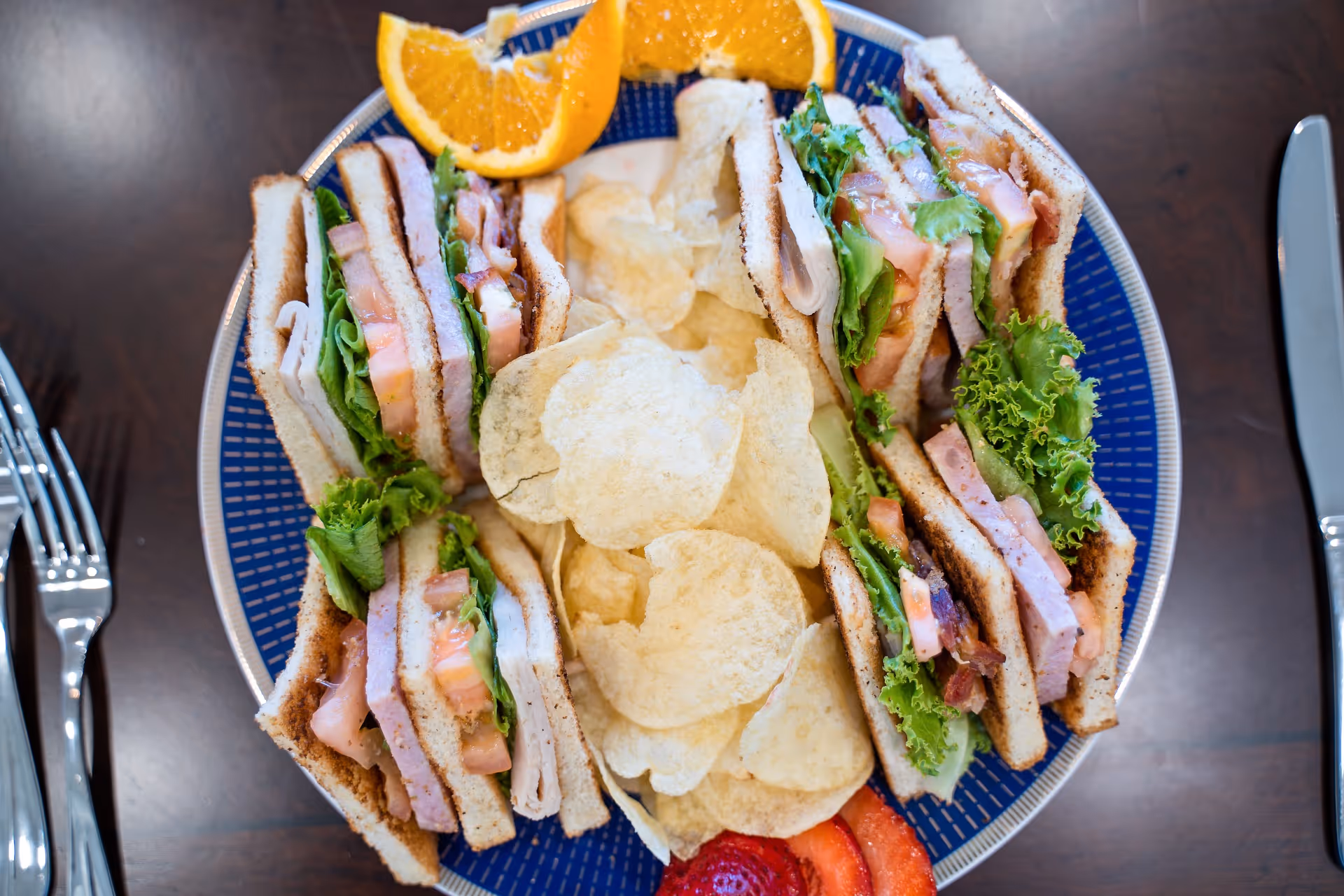 A plate with four club sandwich quarters filled with lettuce, tomato, turkey, and bacon, accompanied by potato chips, orange slices, and strawberry slices on a wooden table with a fork and knife on either side.