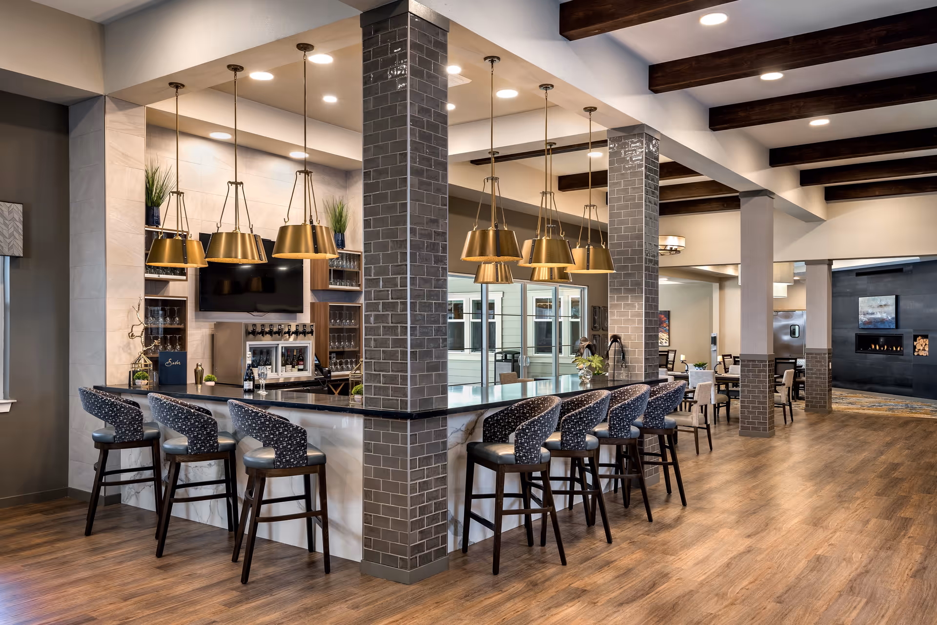 A modern and spacious common area with a large bar counter surrounded by high chairs. The bar features hanging gold pendant lights, a TV mounted on the wall, and shelves with glassware. The room has wooden flooring, exposed dark wooden ceiling beams, and a cozy fireplace in the background with seating areas and tables.
