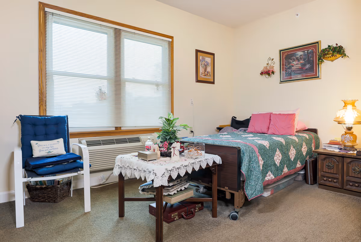 A cozy bedroom with a single bed covered by a quilt, a nightstand with a lamp, a chair by a large window, and a small table with decorations.