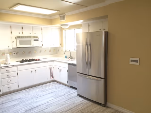 Bright kitchen with white cabinets, a stainless steel refrigerator, a built-in microwave above a gas stove, a dishwasher, and a window above the sink. The floor has light-colored tiles and the walls are painted beige.