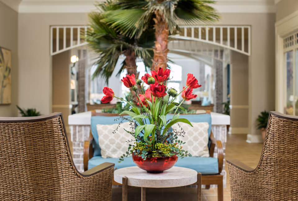 A cozy seating area with two wicker chairs and a blue cushioned loveseat with patterned pillows. A round table in the center holds a red vase with vibrant red flowers and green foliage. In the background, there are tall indoor palm trees and a bright, airy interior space with archways and neutral-colored walls.