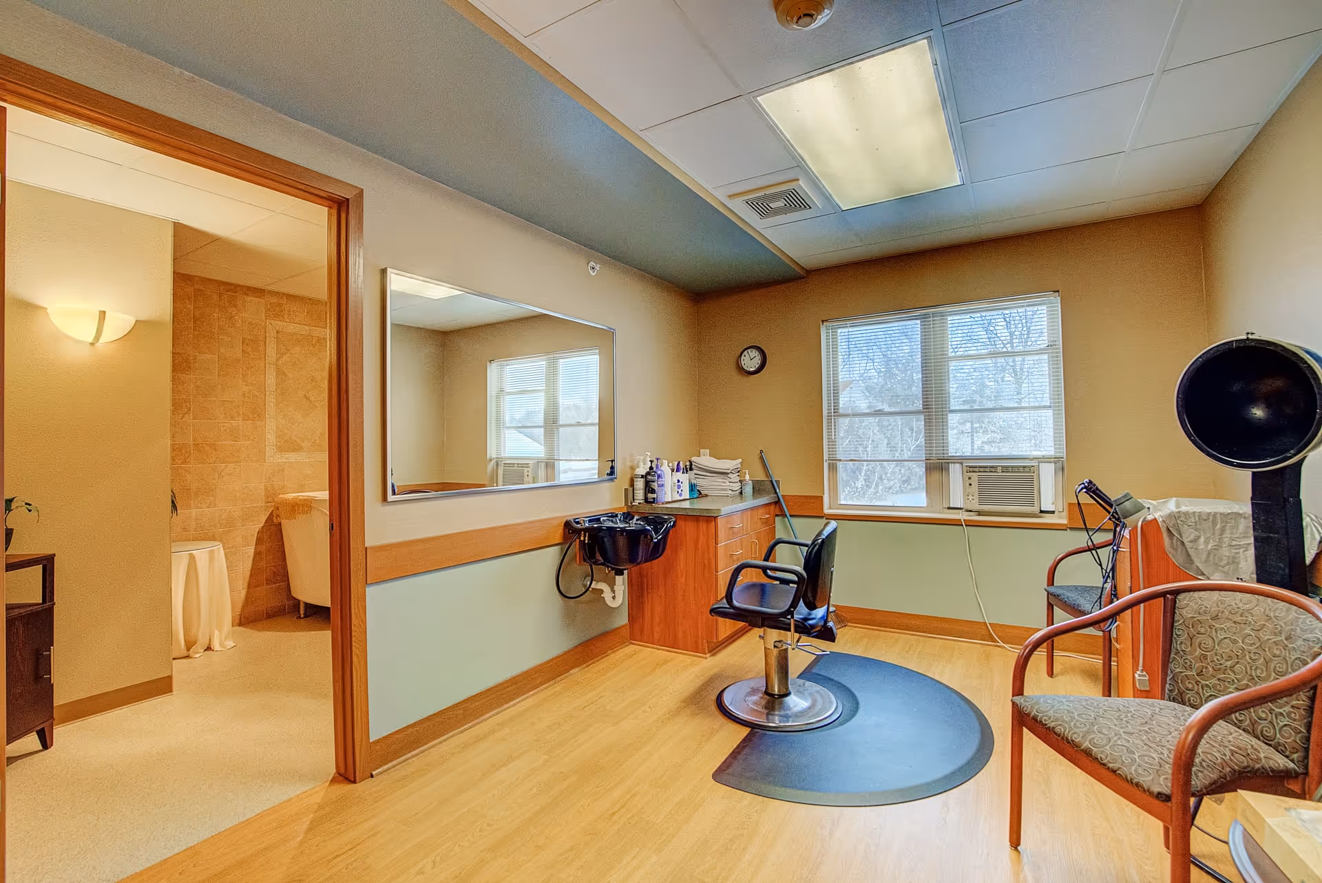 Interior of a salon room in a senior living facility with a black salon chair on a mat, a large mirror on the wall, a countertop with hair care products, and a window with an air conditioning unit. There is also a waiting chair and a hair dryer hood. An adjacent room with a bathtub is visible through an open door.