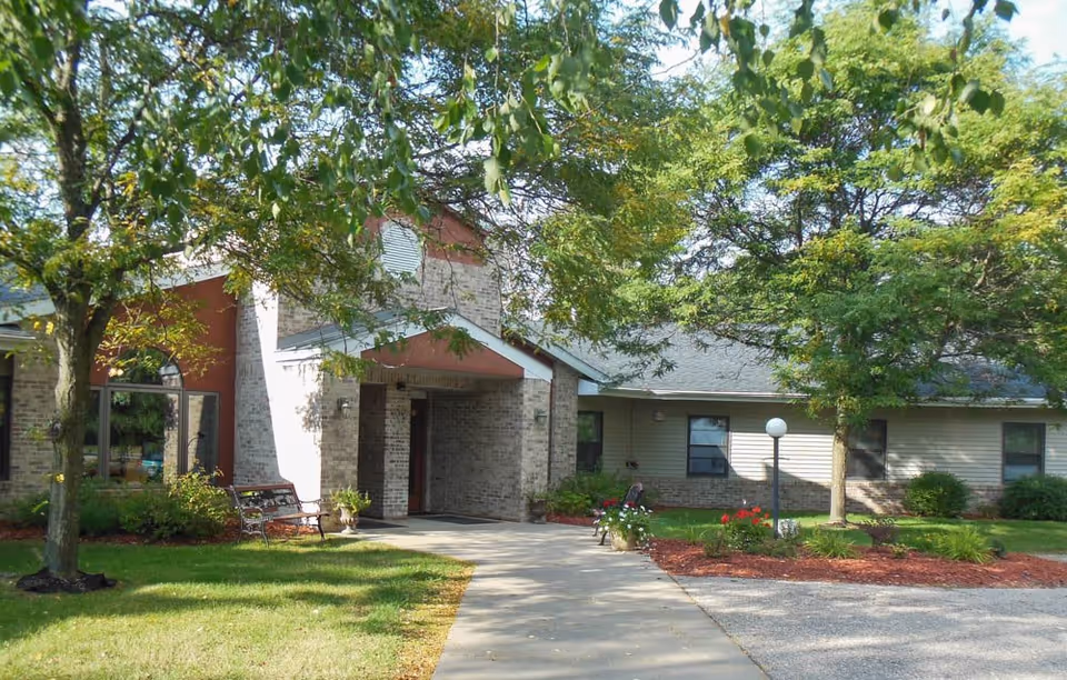 Exterior view of Sylvan Crossings of Stevens Point facility showing a brick and siding building with a covered entrance, surrounded by green trees, shrubs, and a well-maintained lawn with a concrete walkway leading to the entrance.