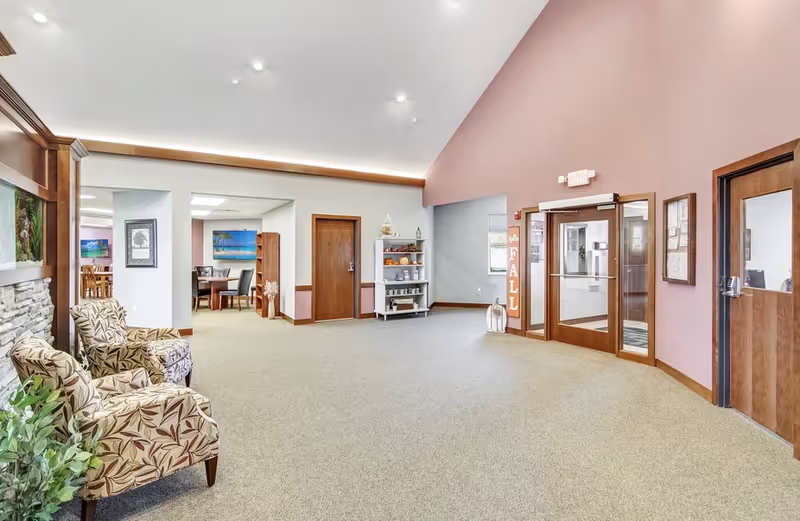 Spacious senior living facility lobby with patterned armchairs, carpeted floor, shelving and glass entry doors.
