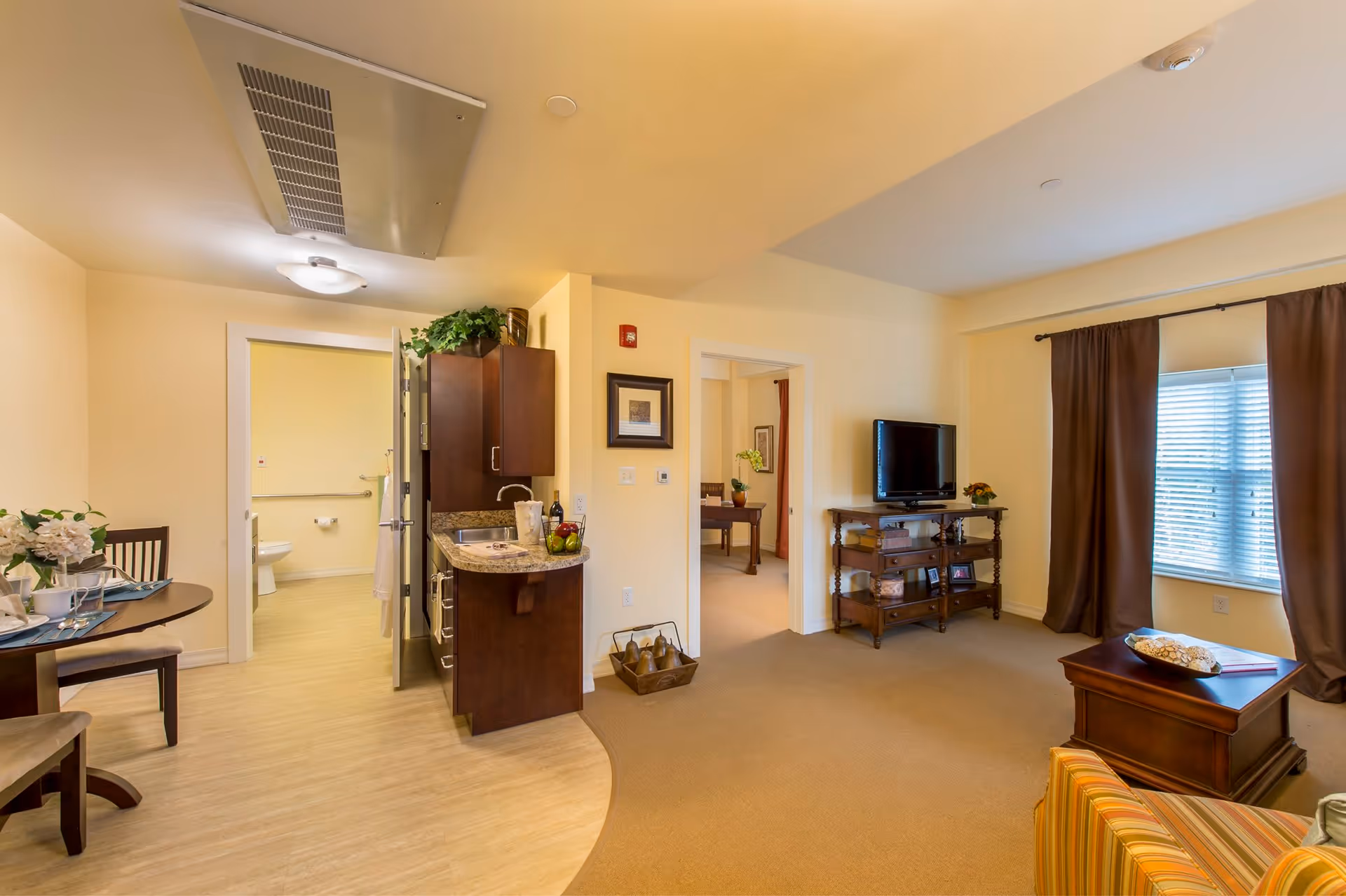 Interior view of a senior living facility apartment showing a living room with a TV on a wooden stand, a coffee table, and a striped armchair. Adjacent to the living room is a small kitchen area with dark wood cabinets and a countertop. A dining table with chairs is visible to the left, and a bathroom with a toilet and grab bars is seen through an open door. Another room is visible through a doorway in the background.