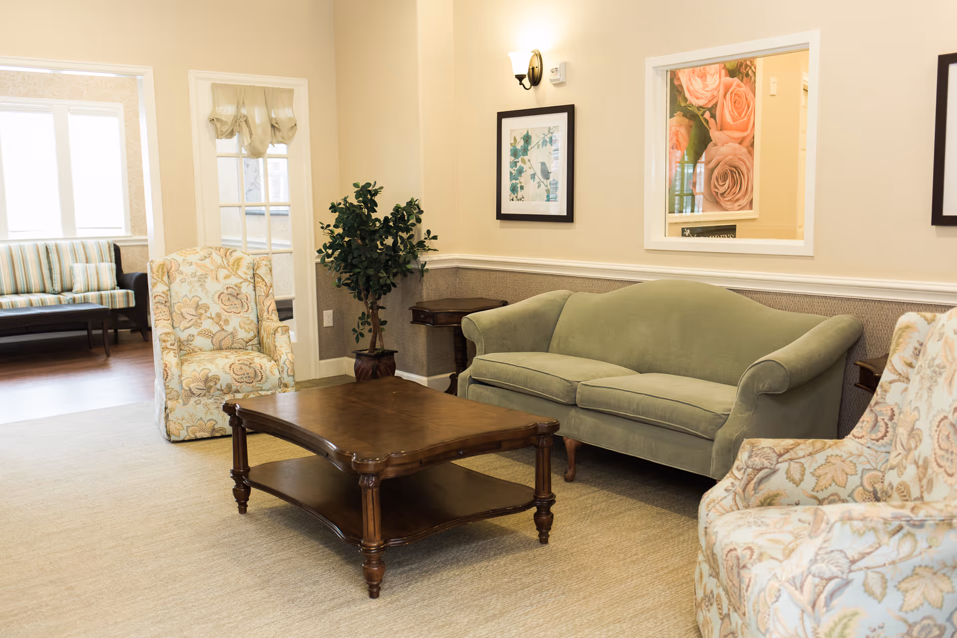 A cozy living room area with a green upholstered sofa, two floral patterned armchairs, a wooden coffee table, a potted plant in the corner, framed artwork on the walls, and a windowed door leading to another room with a striped bench.