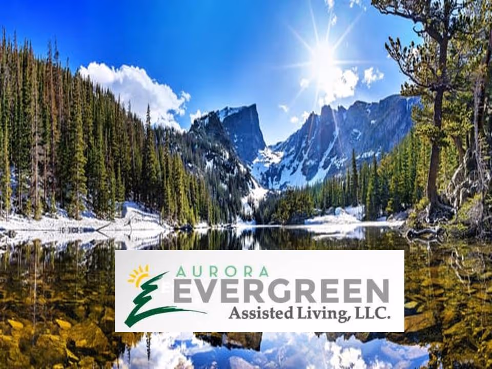 A scenic outdoor landscape featuring a clear lake reflecting surrounding evergreen trees and snow-capped mountains under a bright blue sky with the sun shining.