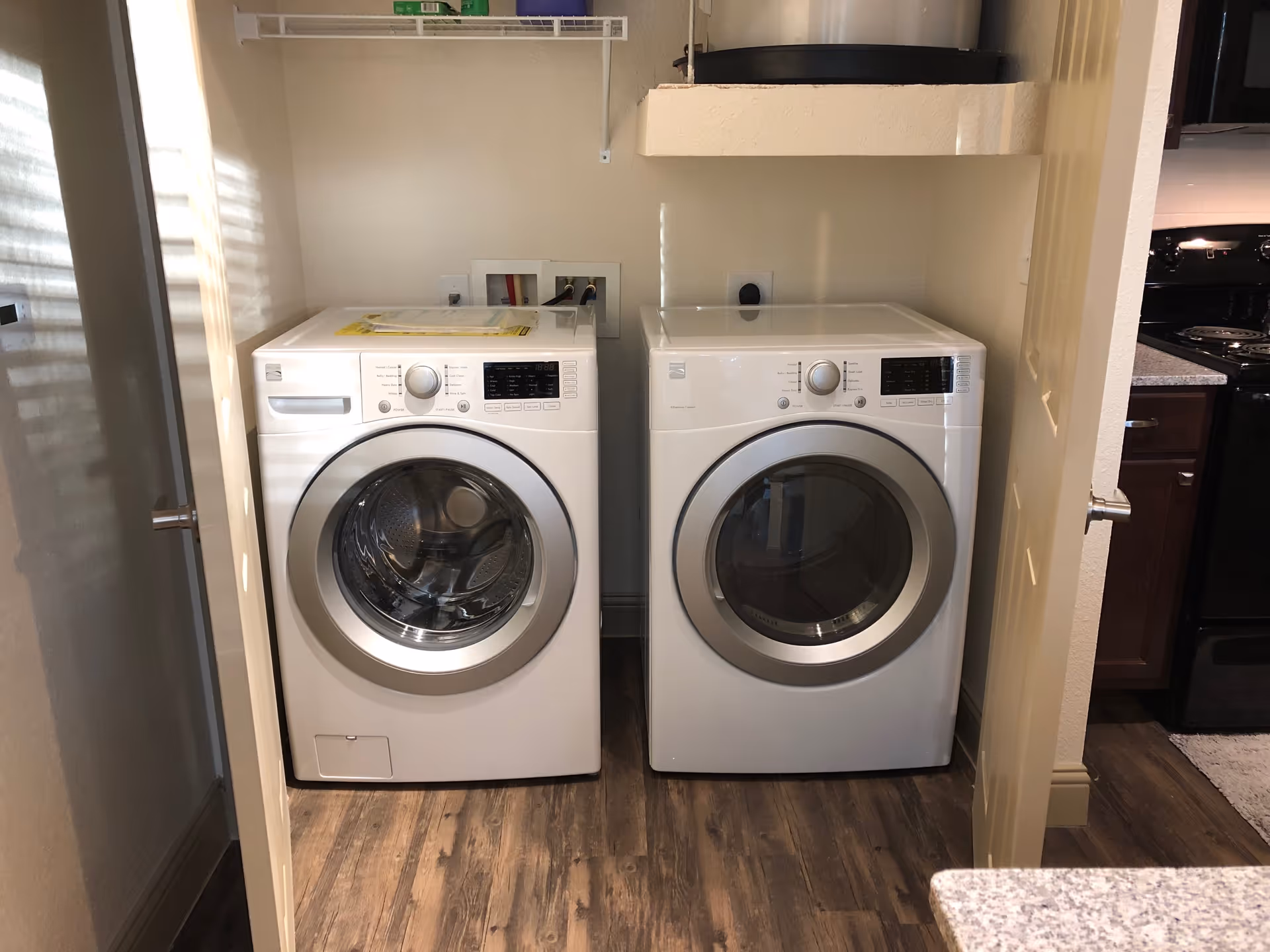 A laundry area with a front-loading washing machine and dryer side by side on a wooden floor. The machines are white and placed in a small nook with beige walls. A shelf is mounted above the machines, and part of a kitchen with dark cabinets and a black stove is visible to the right.