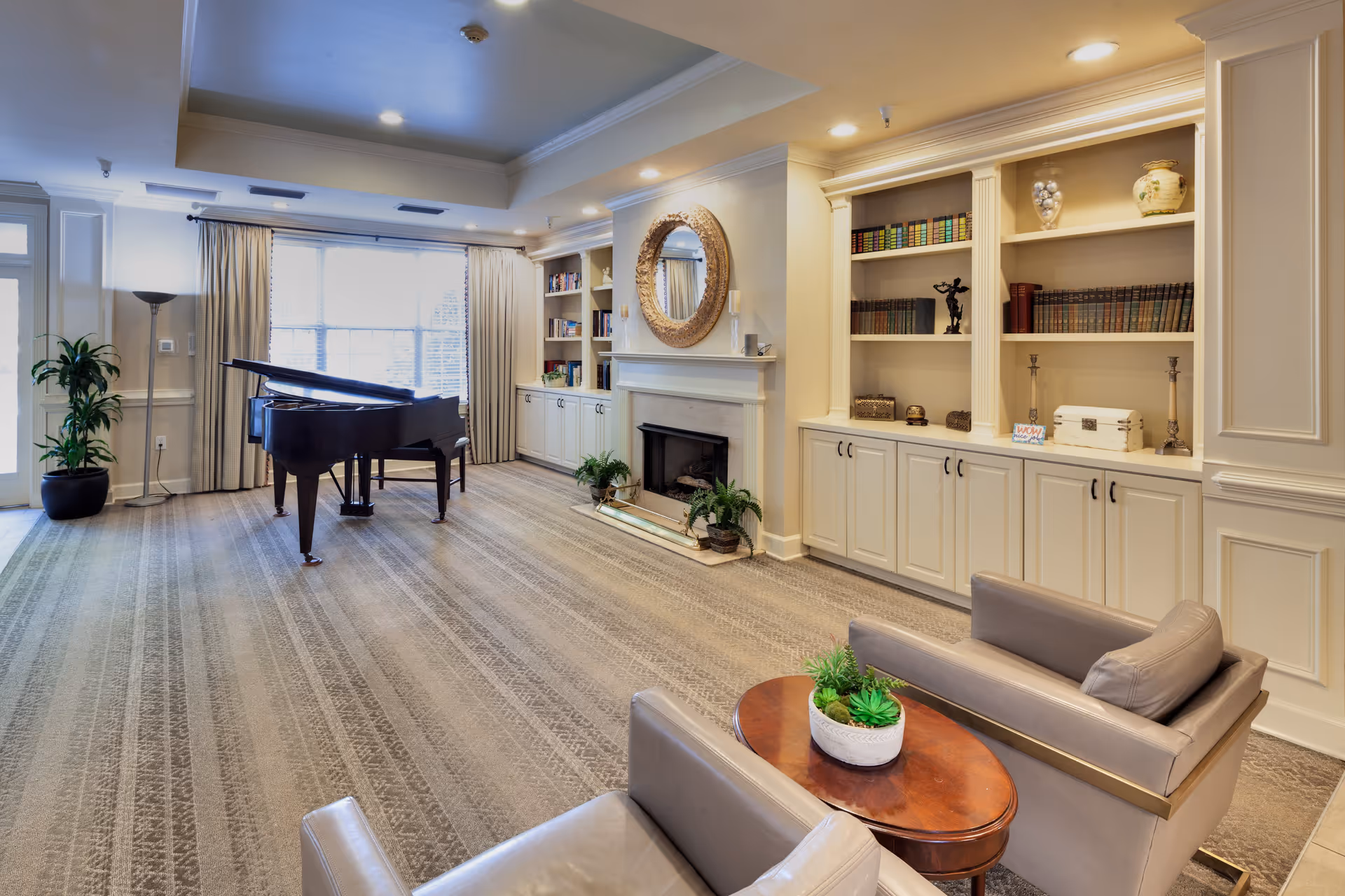 A spacious and well-lit living room with a grand piano near large windows covered with curtains. The room features built-in bookshelves filled with books and decorative items, a fireplace with a round mirror above it, two beige armchairs, and a small wooden table with a potted plant. The carpet has a patterned design, and there are additional plants placed around the room.