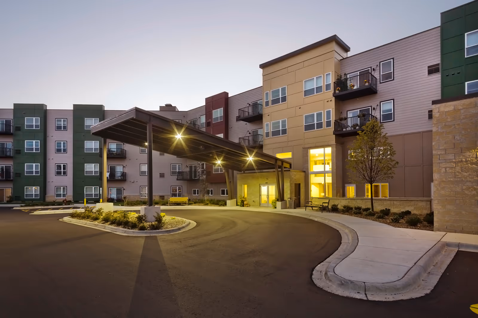 Exterior view of a modern multi-story senior living facility named Woodland Hill at dusk, featuring a covered entrance with lights, balconies, windows, and a curved driveway with landscaping and benches.