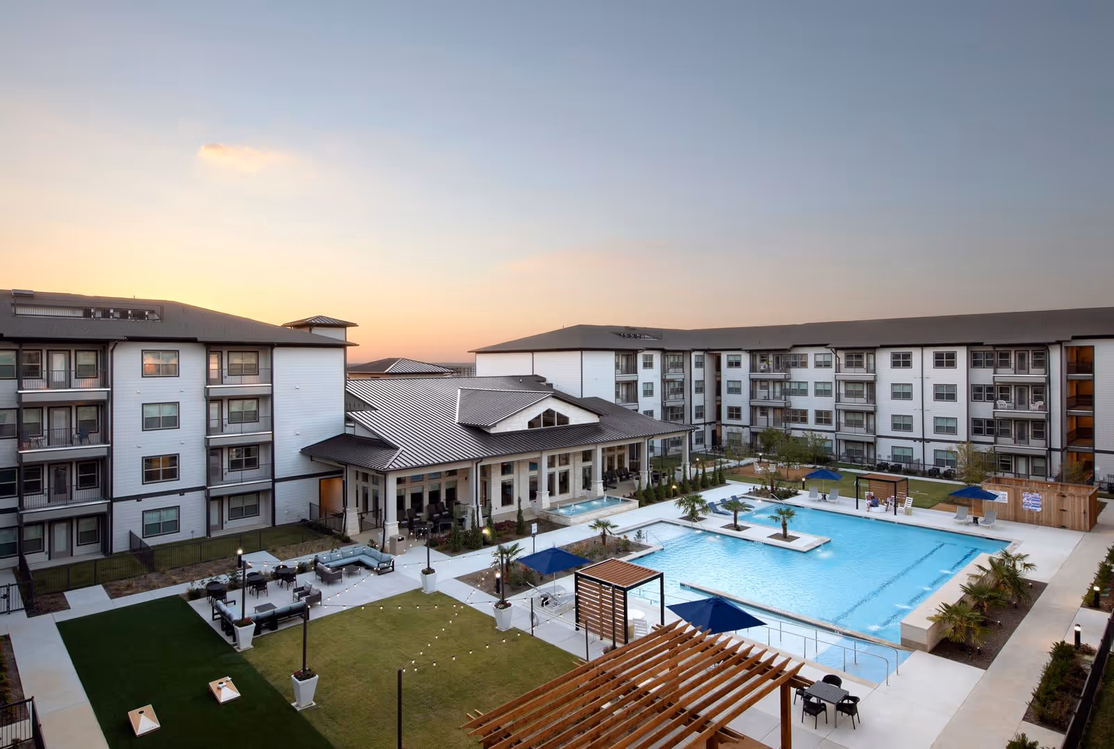 View of an outdoor courtyard area at Solea Tavolo Park featuring a large swimming pool with palm trees on small islands, surrounded by lounge chairs and umbrellas. The courtyard is enclosed by a multi-story residential building with balconies. There is a covered seating area, a pergola, and a grassy area with a cornhole game set up. The sky is clear with a soft sunset glow.