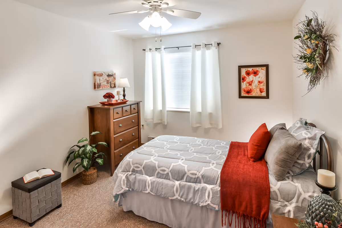 A cozy bedroom with a neatly made bed featuring gray and white patterned bedding and a red throw blanket. The room has a wooden dresser with a lamp and decorative items on top, a window with white curtains, a small plant in a woven basket, a framed floral picture on the wall, and a decorative wreath. The walls are painted white and the floor is carpeted.