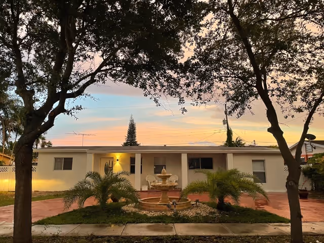 Single-story house front with a central tiered fountain, small palm plants, red driveway and trees framing a colorful sunset sky.
