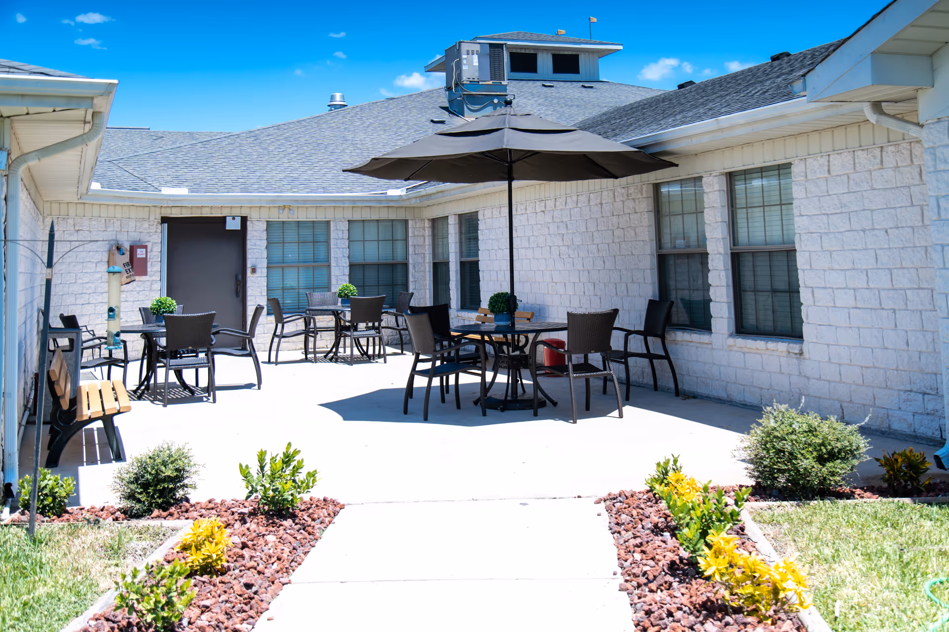 Sunny courtyard patio with tables, chairs, an umbrella and small landscaped beds outside a nursing facility.