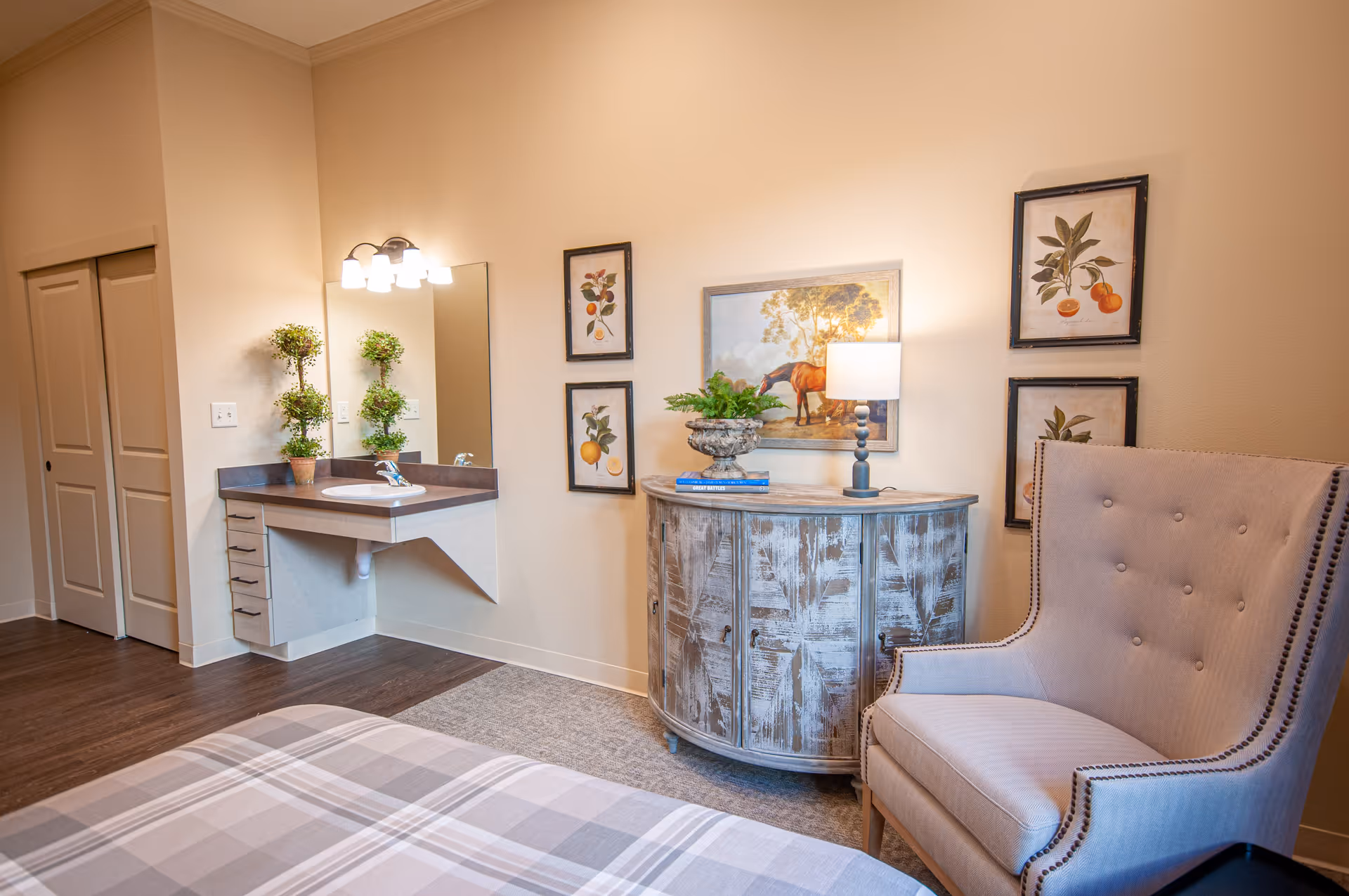 A cozy senior living bedroom featuring a plaid bedspread in the foreground, a beige upholstered armchair with button tufting and nailhead trim, a rustic wooden cabinet with a lamp and plant on top, and framed botanical and horse artwork on the walls. A corner sink with a mirror and potted plant is also visible, along with a closet with sliding doors.
