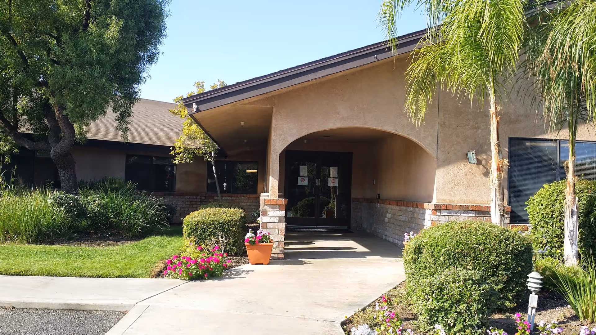 Entrance to a single-story building with an arched covered walkway, potted flowers and landscaped shrubs and palm trees.
