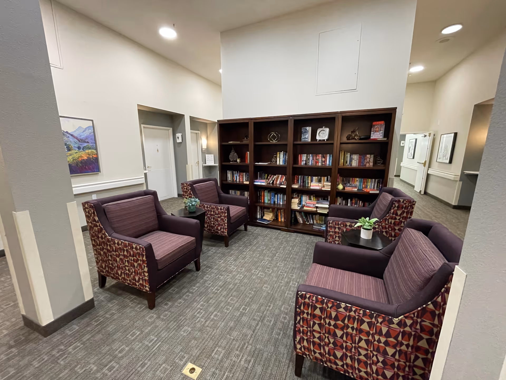 Four purple patterned armchairs arranged around side tables facing a large bookshelf filled with books in a senior living facility common area.