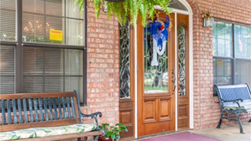 Front entrance of a building with a wooden door featuring decorative glass panels, flanked by two large windows with blinds. There are two benches with cushions on either side of the door, a hanging fern plant above, and a small potted plant near one bench. The exterior wall is made of red brick.