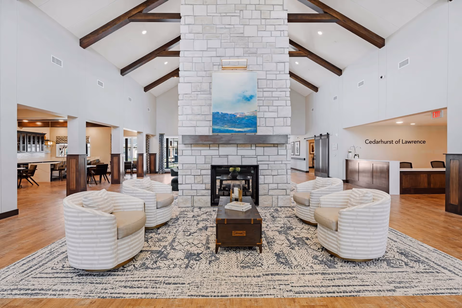 A spacious senior living facility common area with a high vaulted ceiling featuring exposed wooden beams. In the center is a large stone fireplace with a blue and white painting above it. Surrounding the fireplace are six cushioned armchairs arranged in a semi-circle on a patterned area rug. To the left, there is a dining area with tables and chairs, and to the right is a reception desk with the text 'Cedarhurst of Lawrence' on the wall behind it. The flooring is wood, and the space is well-lit with recessed lighting.
