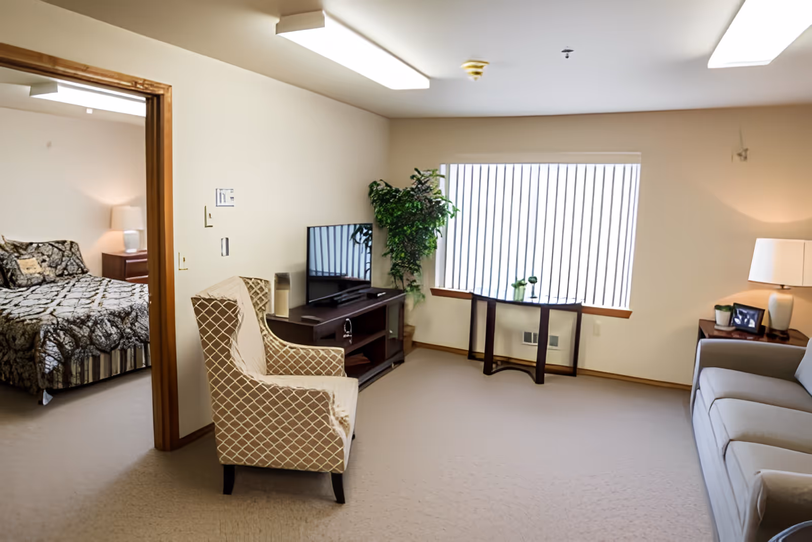 A bright living room with a beige carpet, a patterned armchair, a gray sofa, a wooden TV stand with a flat-screen TV, a small table under a large window with vertical blinds, a potted plant, and a side table with a lamp and framed photo. An adjacent bedroom with a bed covered in a patterned bedspread and a nightstand with a lamp is visible through an open doorway.