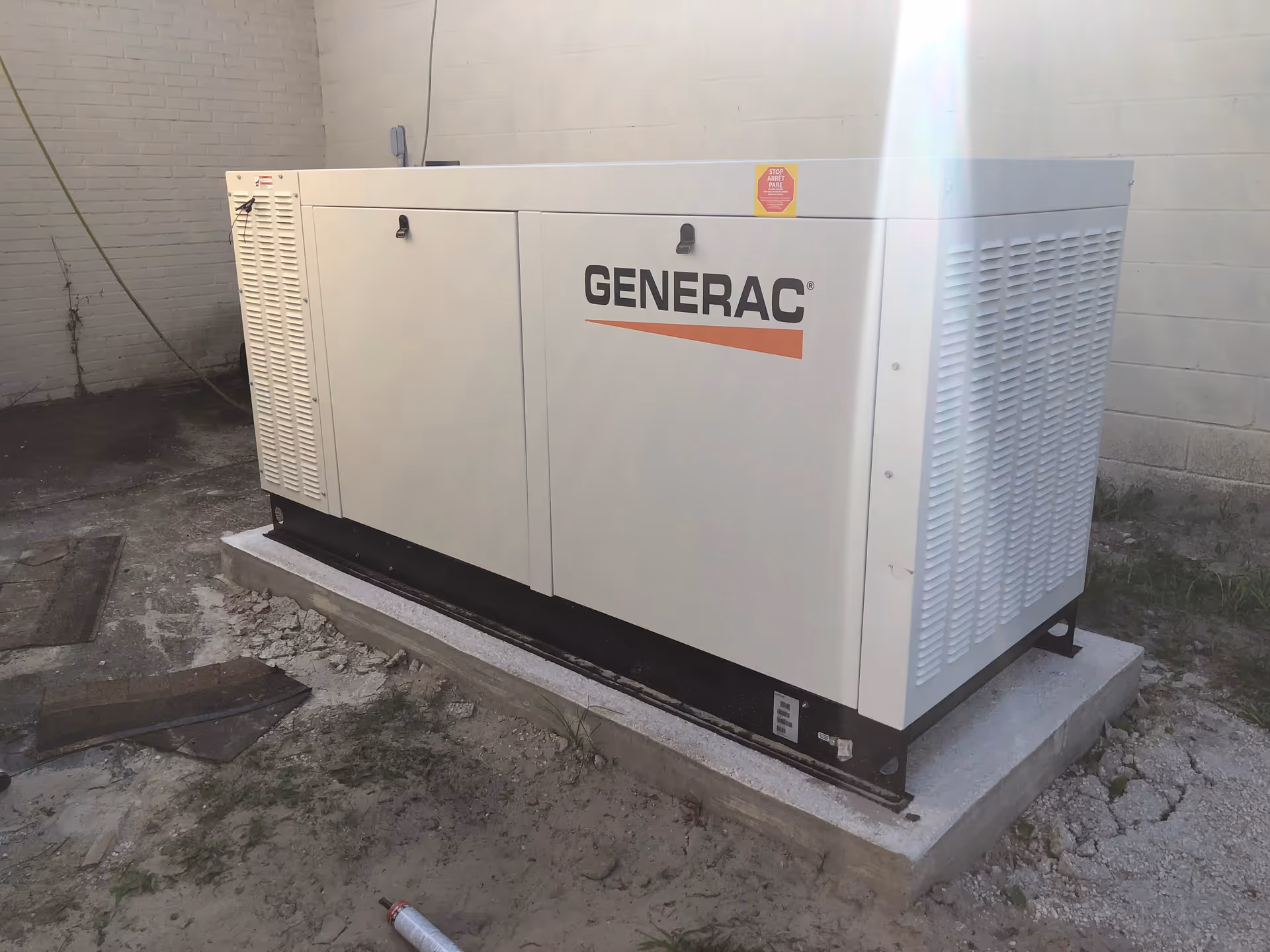 A large white Generac generator installed on a concrete platform outside next to a white brick wall with some dirt and construction debris around it.