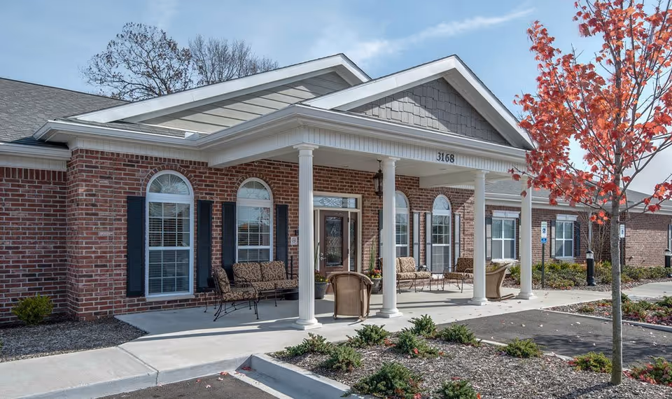 Front exterior view of a single-story brick building with white columns supporting a covered entrance. There are several windows with black shutters and outdoor seating with cushioned chairs and a small table on the porch. A tree with red autumn leaves is visible on the right side near the parking area.