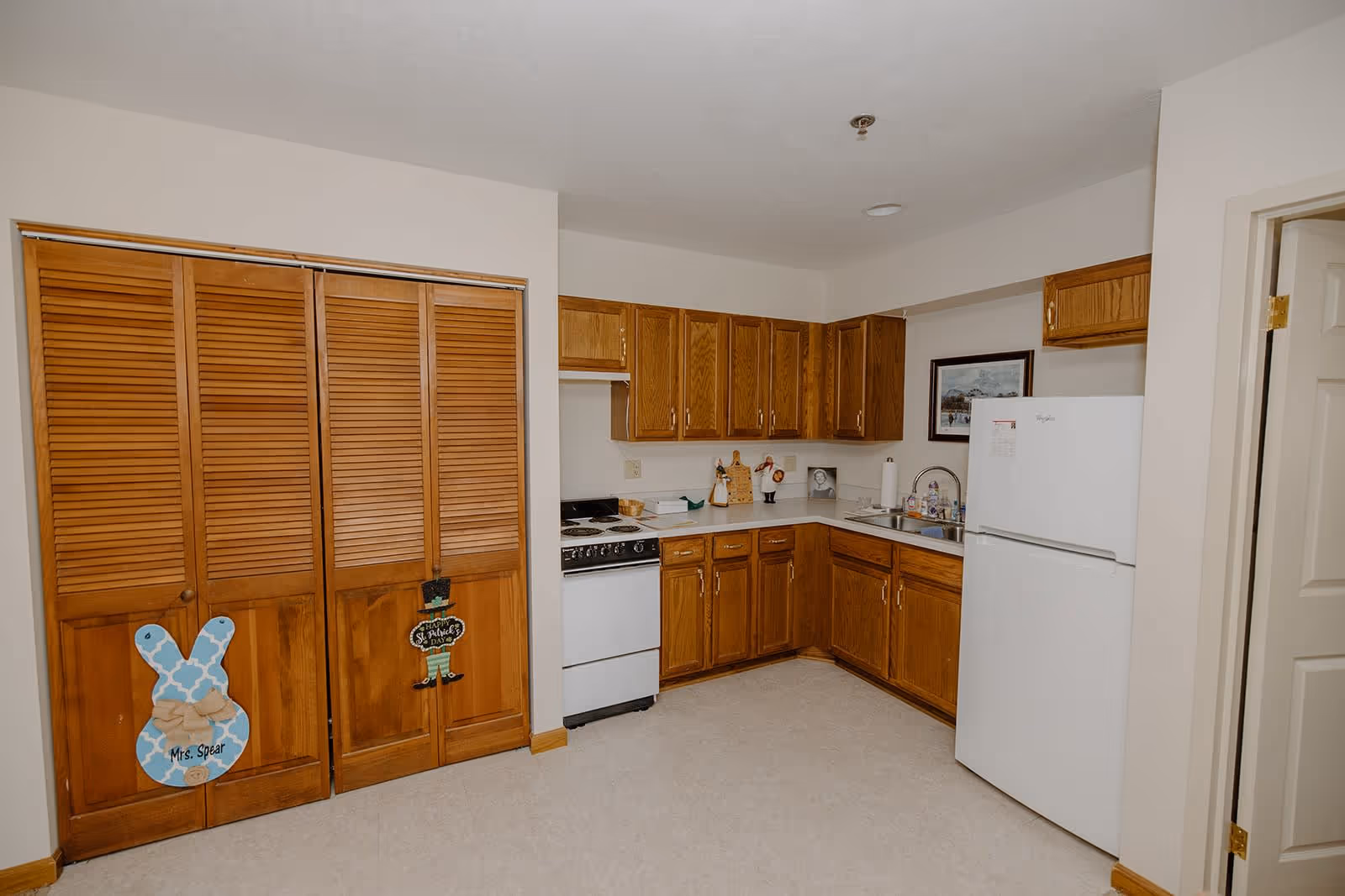 Interior view of a kitchen area in an assisted living facility featuring wooden cabinets, a white refrigerator, a stove, a double sink, and a set of wooden closet doors with decorative signs attached.