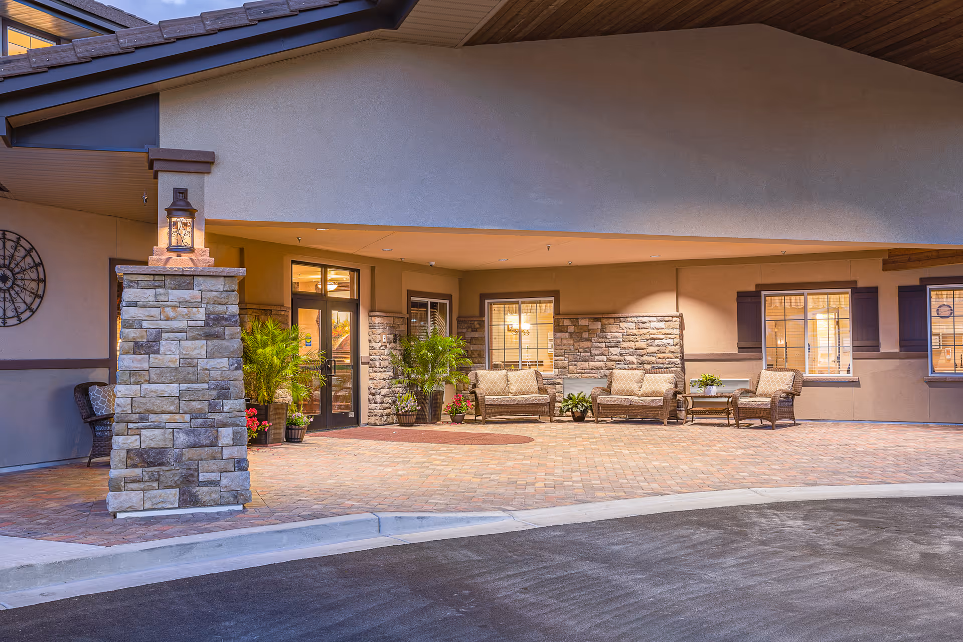 Entrance area of a senior living facility with stone pillars, potted plants, and outdoor seating including cushioned chairs and a table. The building exterior features stone and stucco walls with windows and warm lighting inside.