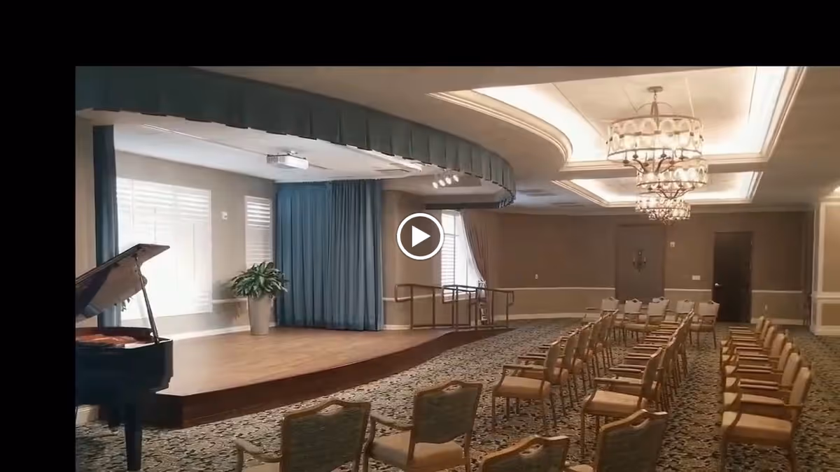 A large interior event room with a small stage and piano at left, rows of chairs facing the stage, and chandeliers overhead.