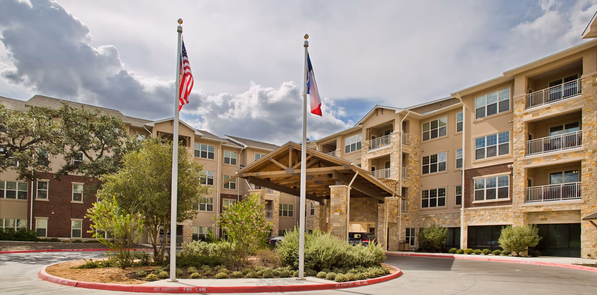 Exterior view of a senior living facility with a covered entrance supported by stone pillars. Two flagpoles with the American flag and another flag stand in front of the entrance. The building is multi-story with balconies and a mix of stone and brick facade. There are trees and shrubs in landscaped areas around the driveway.