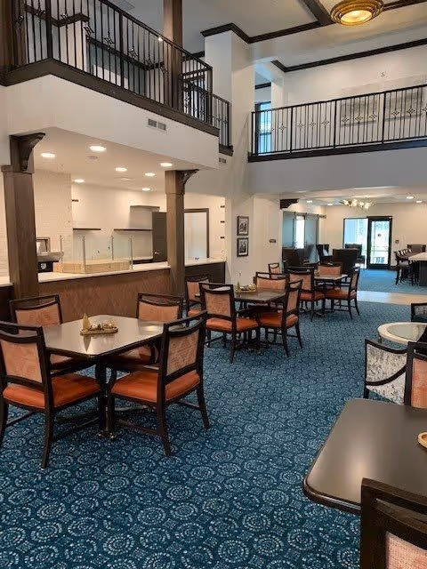 Interior view of a senior living facility dining area with multiple tables and chairs arranged on a blue patterned carpet. The space features high ceilings with a balcony railing above, warm lighting, and a kitchen area visible in the background.