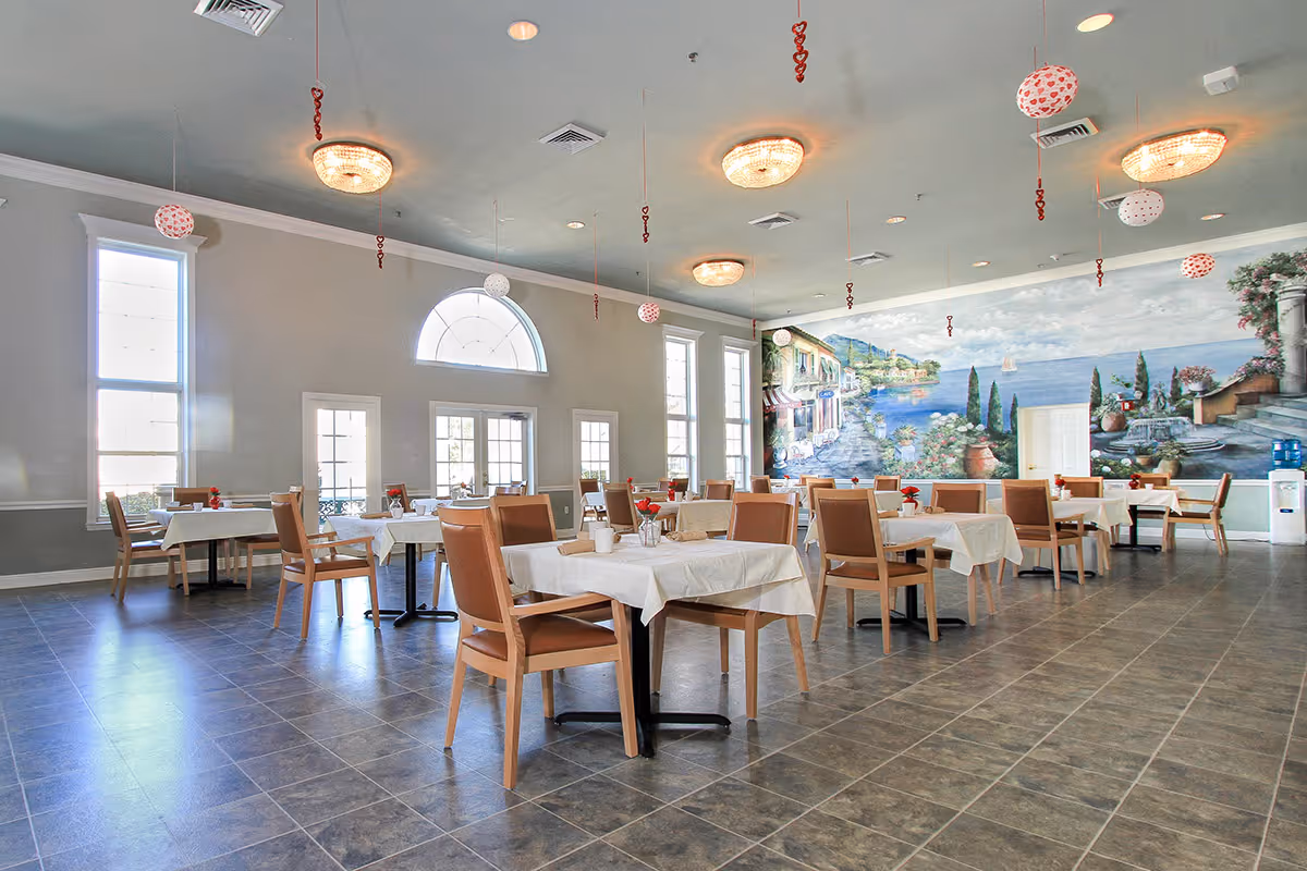 A spacious dining room with multiple tables covered in white tablecloths and surrounded by wooden chairs with tan cushions. The room features large windows allowing natural light to fill the space, and a mural on one wall depicting a scenic coastal village with water, greenery, and buildings. Ceiling lights and hanging decorations add to the ambiance.