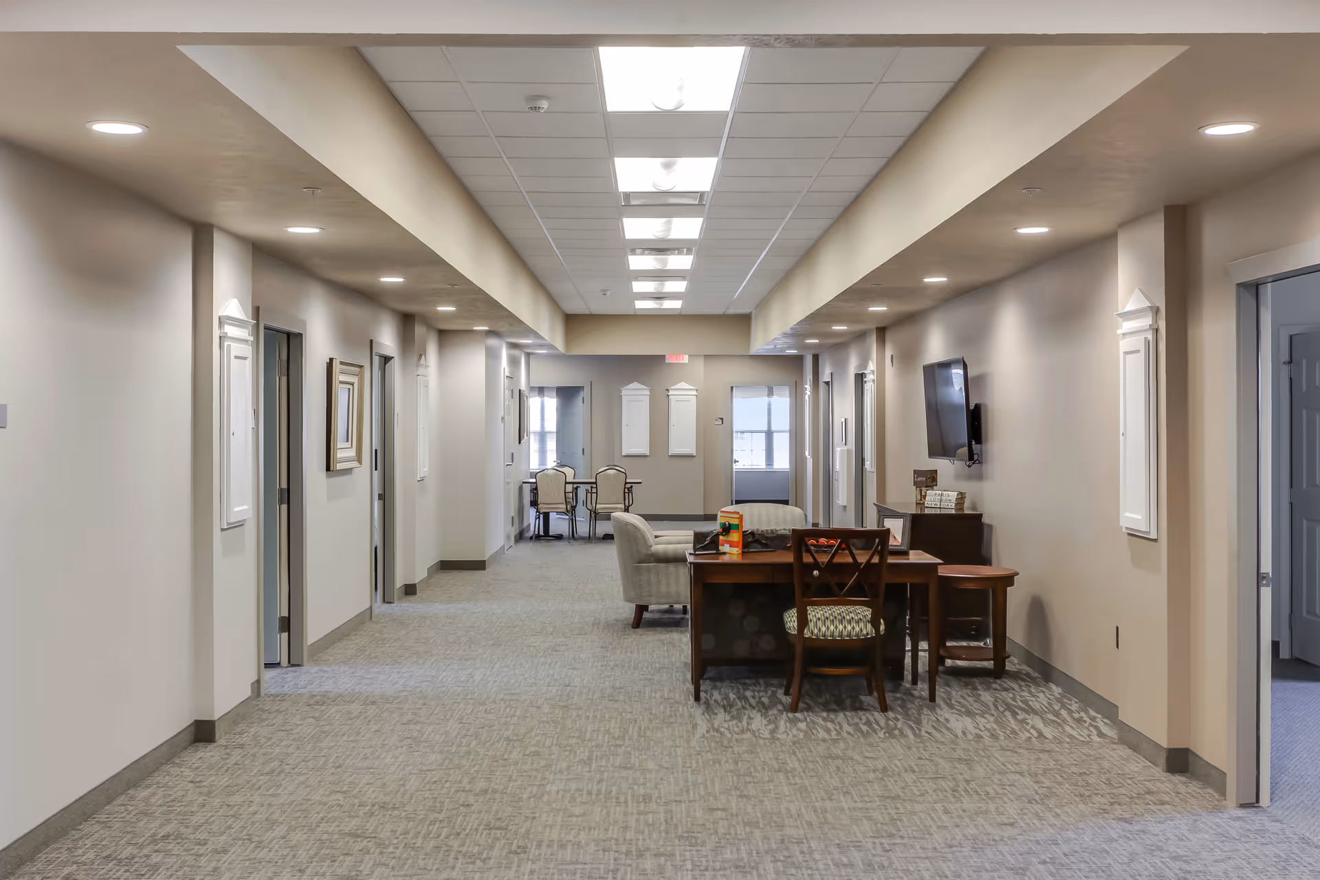 A wide hallway in an assisted living facility with beige walls and carpeted floor. The hallway has several doorways on both sides, a table with chairs and a couch in the middle, and a wall-mounted TV on the right side. The ceiling has recessed lighting and skylights.