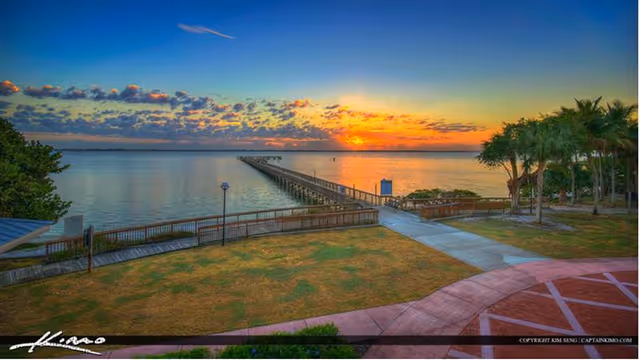 A scenic view of a waterfront at sunset with a long wooden pier extending into the calm water. The sky is partly cloudy with vibrant orange and blue hues. In the foreground, there is a grassy area bordered by a curved walkway and some trees on the right side.