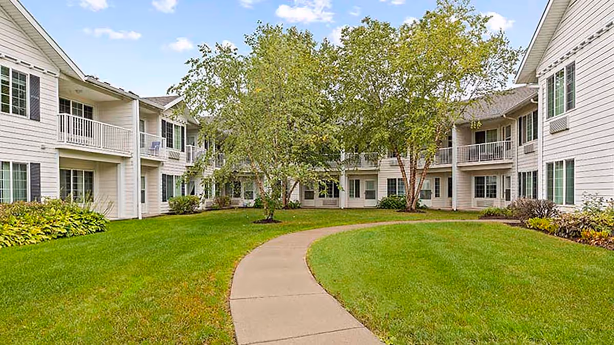 A curved concrete pathway leads through a green lawn with trees and shrubs, surrounded by a two-story white residential building with balconies and windows under a partly cloudy sky.