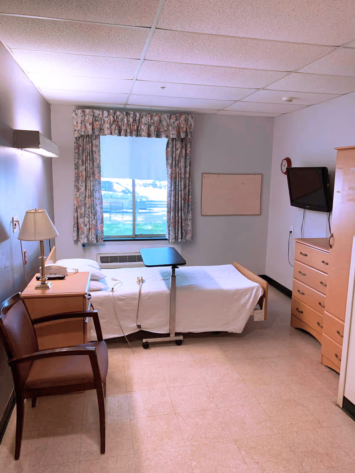 A simple, clean room with a single bed covered in white sheets, a small overbed table, a wooden nightstand with a lamp and telephone, a wooden chair, a window with floral curtains, a wall-mounted TV, a wooden dresser, and a clock on the wall.