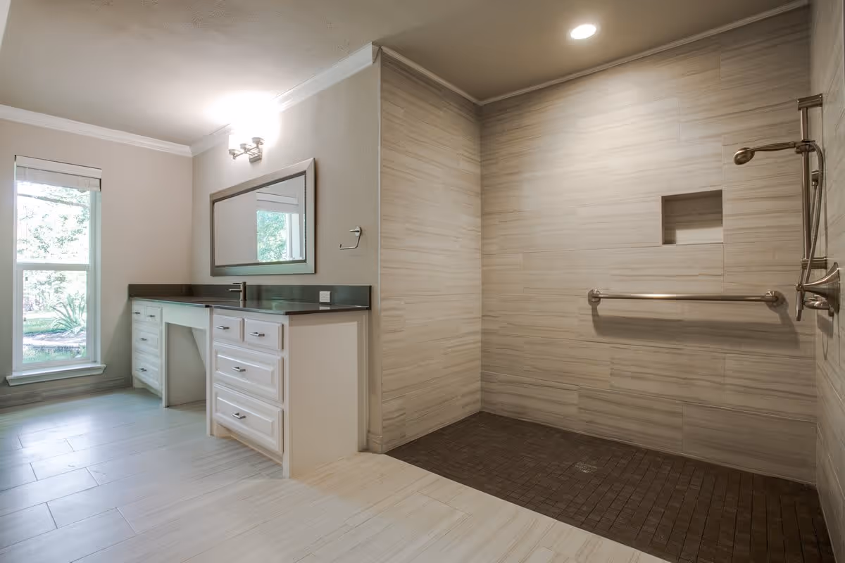 A spacious bathroom featuring a large walk-in shower with beige tiles and a built-in niche, equipped with a handheld showerhead and a grab bar. To the left, there is a vanity with a dark countertop, white cabinets, a large mirror, and a window letting in natural light.