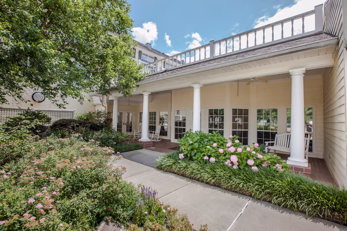 Covered entrance porch of a senior living building with white columns, benches, and landscaped flower beds.