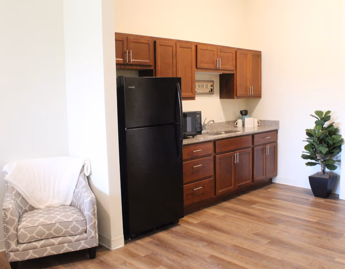 A small kitchen area with dark wooden cabinets, a black refrigerator, a microwave, a sink, and a granite countertop. To the left of the kitchen is a patterned armchair with a white throw blanket draped over it. On the right side, there is a potted green plant on the wooden floor.