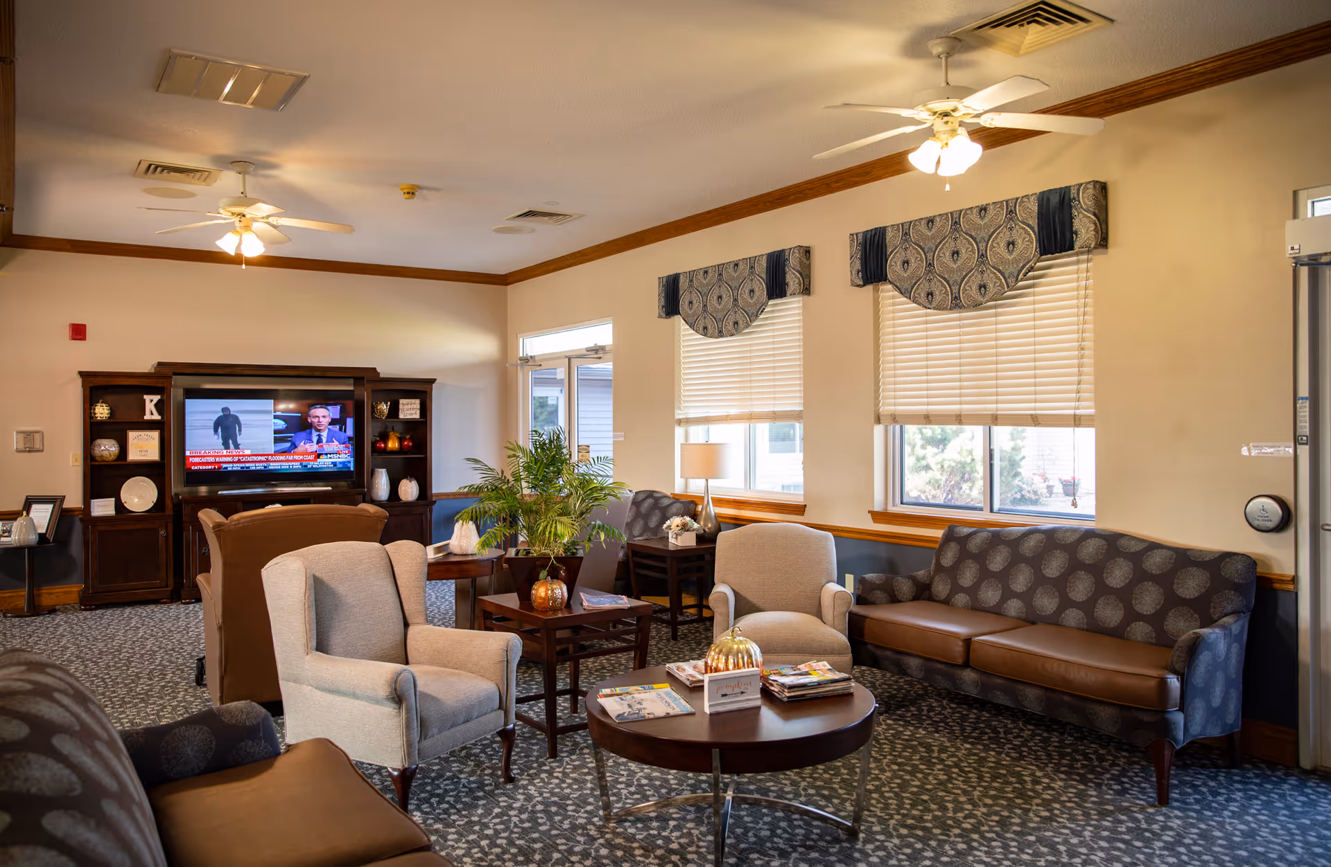 A cozy living room area in an assisted living facility with patterned sofas and armchairs arranged around wooden coffee tables. The room has two windows with blinds and decorative valances, ceiling fans with lights, a TV on a wooden entertainment center, and a potted plant on a side table.