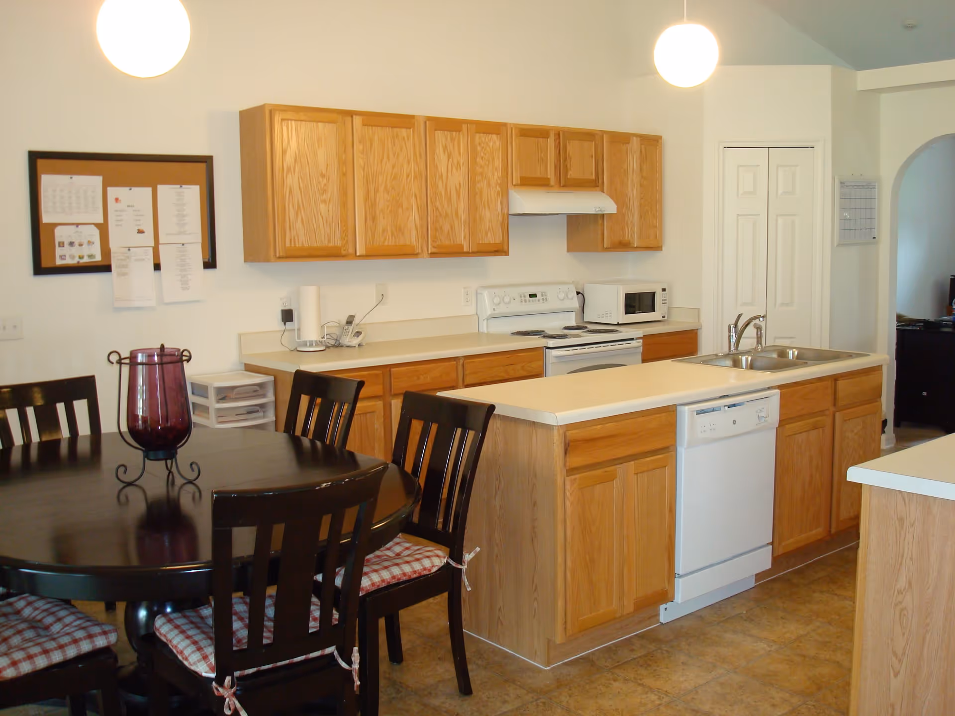 A kitchen with wooden cabinets, a white stove, microwave, dishwasher, and a double sink. There is a round dining table with four chairs that have plaid seat cushions. A purple decorative vase is on the table. The walls are white, and there are two hanging round light fixtures above the kitchen and dining area.