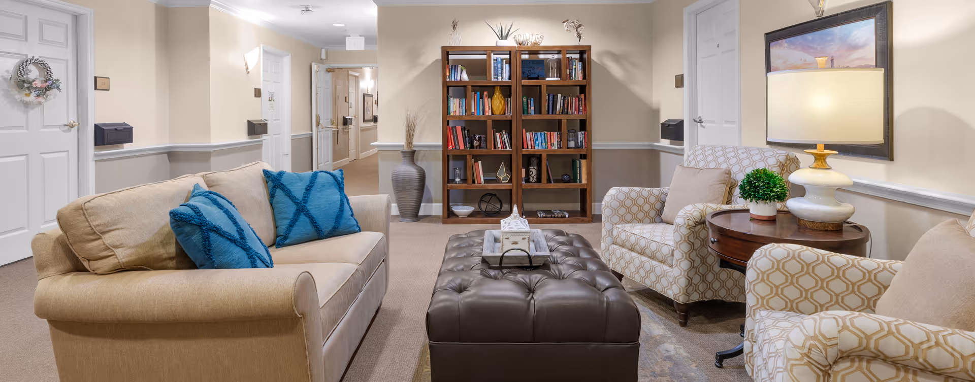 A cozy sitting area in a senior living facility hallway featuring a beige sofa with blue pillows, two patterned armchairs with beige cushions, a large tufted leather ottoman, a wooden bookshelf filled with books and decorative items, a round wooden side table with a lamp and a small plant, and a framed painting on the wall.