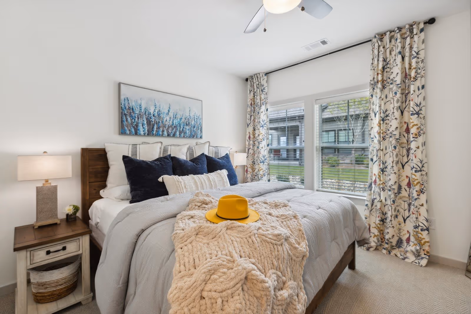 A cozy bedroom with a neatly made bed featuring multiple pillows in white and navy blue, a chunky knit beige throw blanket, and a yellow hat placed on top. The room has a wooden nightstand with a lamp and a small plant, floral patterned curtains, two large windows with blinds, and a ceiling fan with a light.